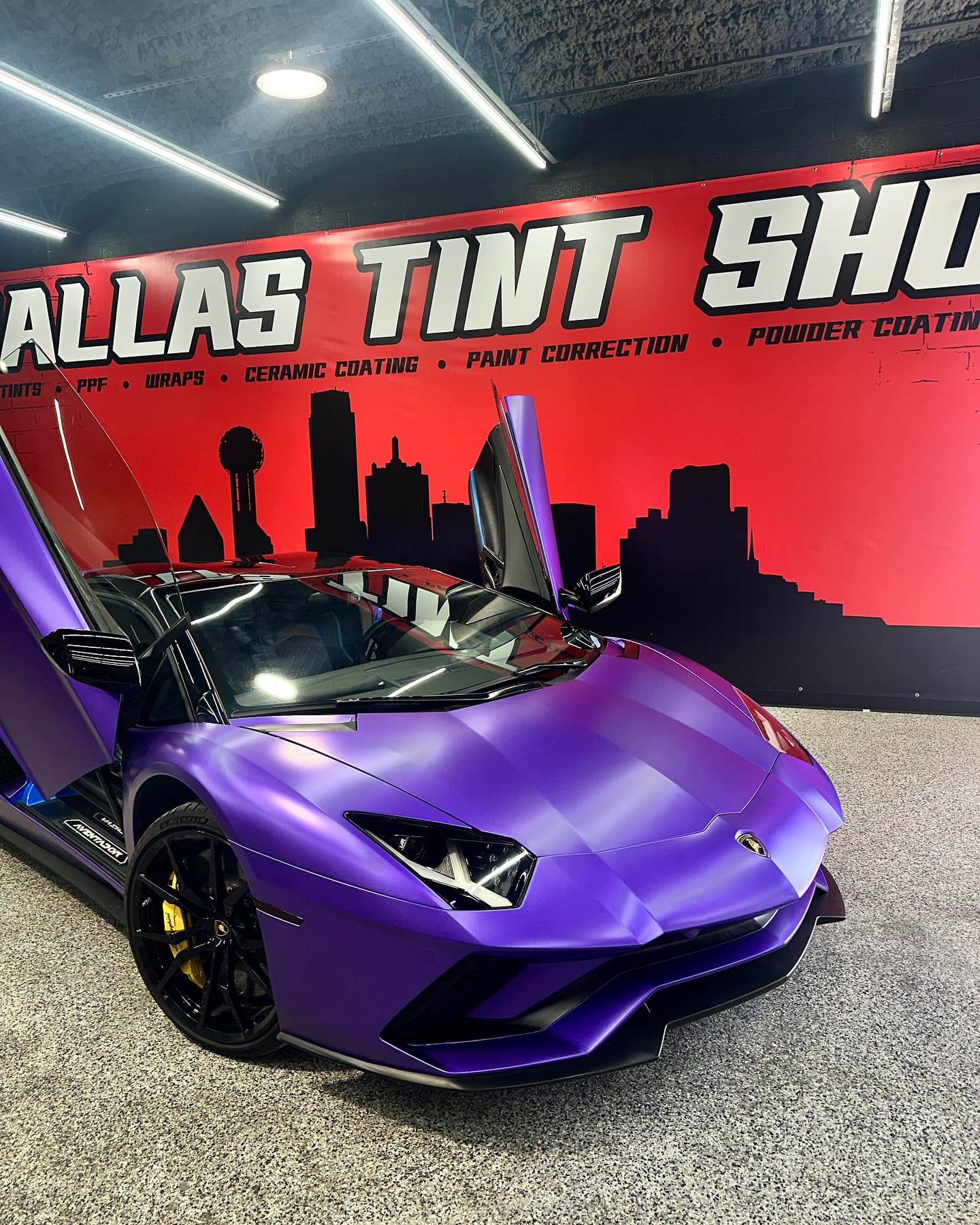 Purple Lamborghini Aventador with the doors up against the Dallas Tint Shop mural